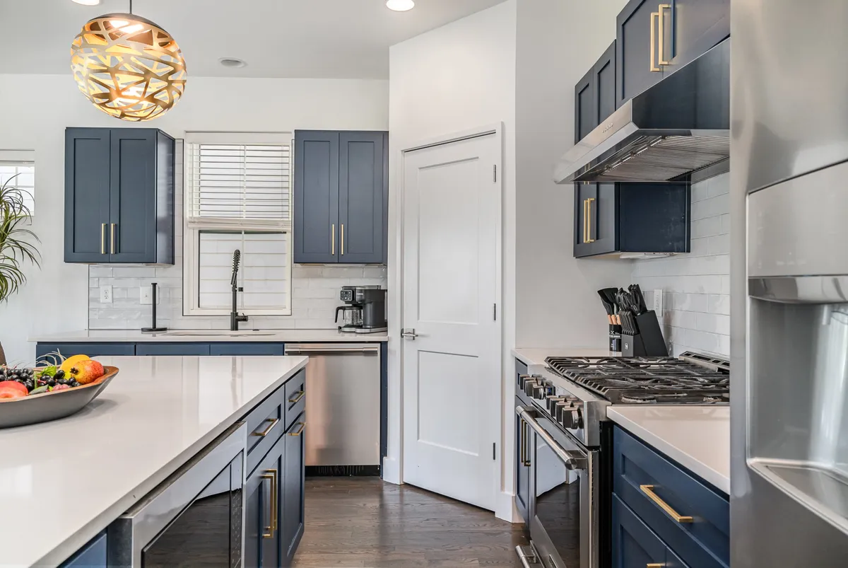 A stylish kitchen with striking blue cabinetry, a marble countertop, gold light fixture, and stainless steel appliances. A fruit bowl on the counter adds a welcoming touch. The kitchen is ready for cooking and entertaining, aligning with the section’s emphasis on shared meals and culinary inspiration.