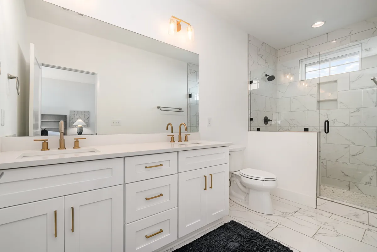 A spacious, spa-inspired bathroom with a marble-tiled shower enclosure, vanity with gold fixtures, and expansive mirrors. The design is bright and inviting, evoking a hotel-style experience.