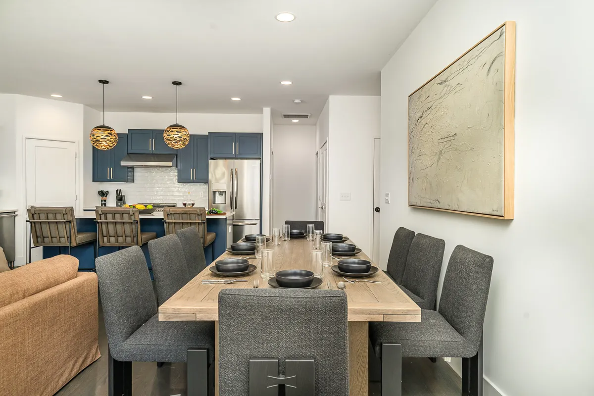 A contemporary dining area with a spacious table, kitchen island in the background, pendant lighting, and artwork on the wall. The scene suggests a setting ready for family dinners, group games, or celebratory toasts, matching the section’s description of a versatile dining space for eight.