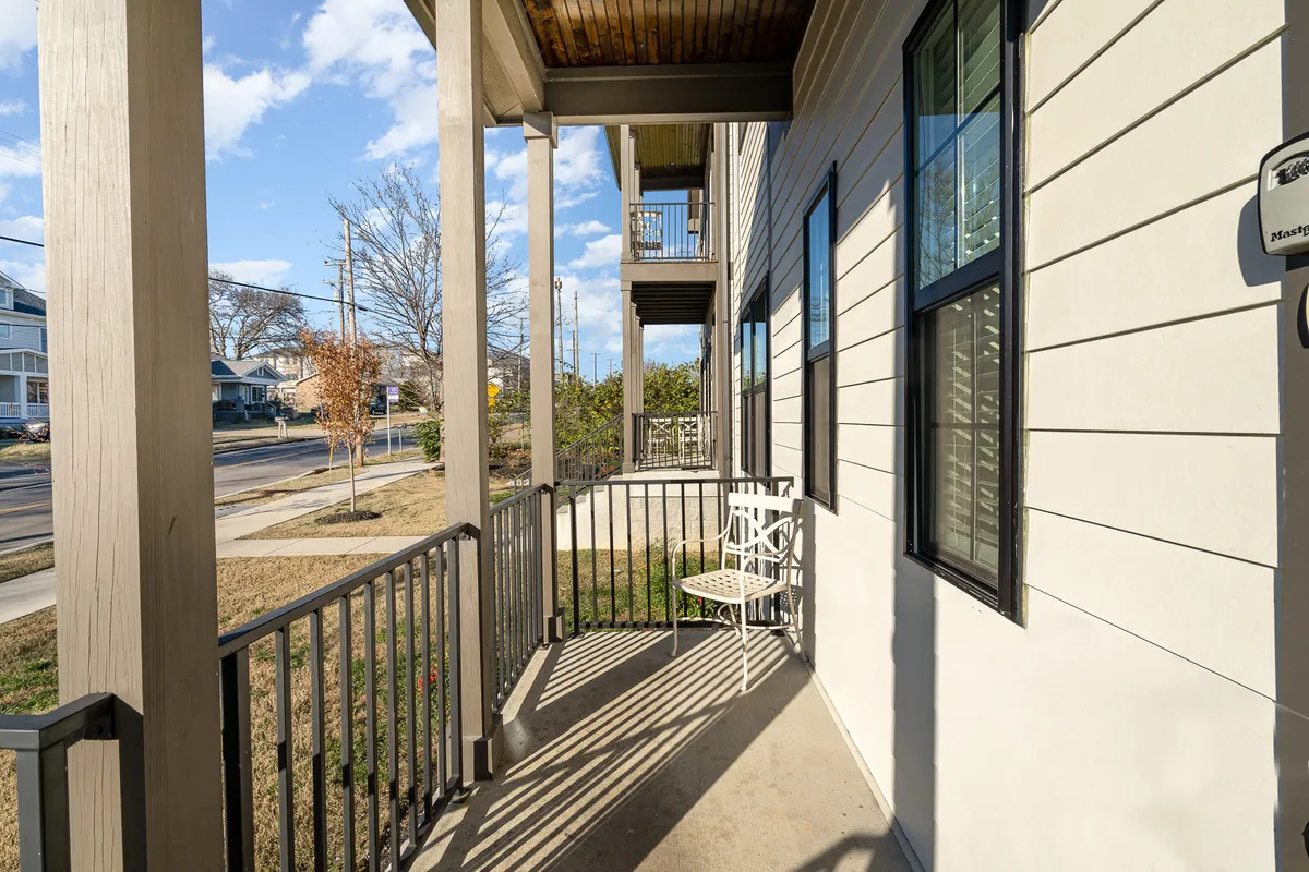 This image captures a private porch area featuring a white chair, porch railing, and the exterior walls of the home, with blue skies and neighboring houses visible in the background. The overall mood is relaxed and inviting, with an emphasis on sitting and enjoying a morning coffee or reading a book in the fresh air.